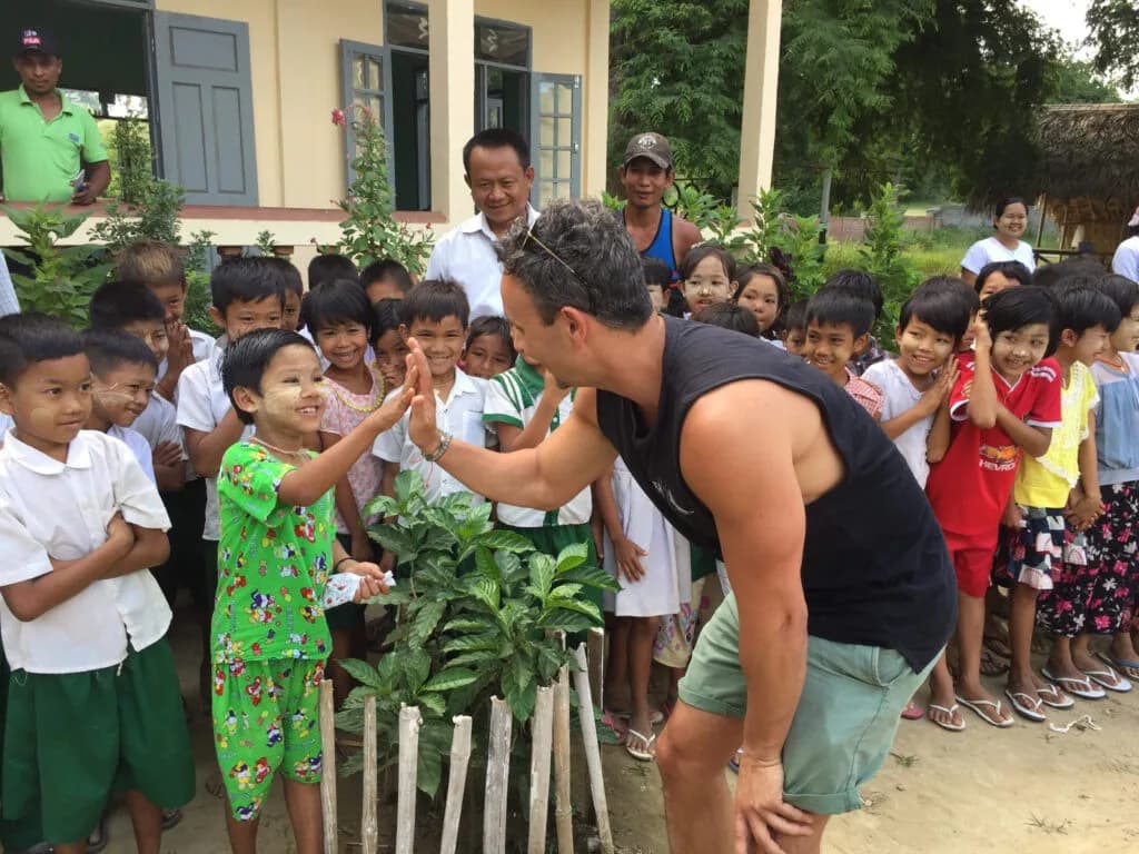 A visitor high-fiving a young student beside a newly planted garden, with a crowd of students watching