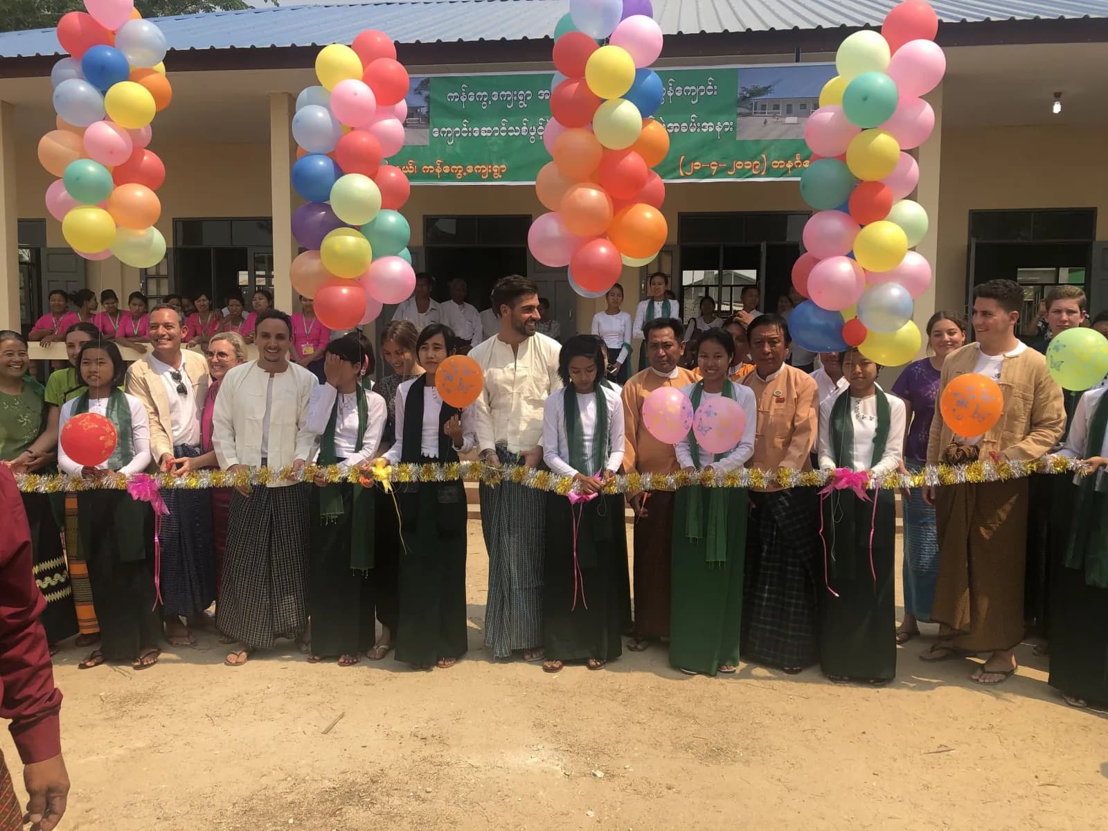 A crowd lined up behind a ribbon outside a school decorated with balloons