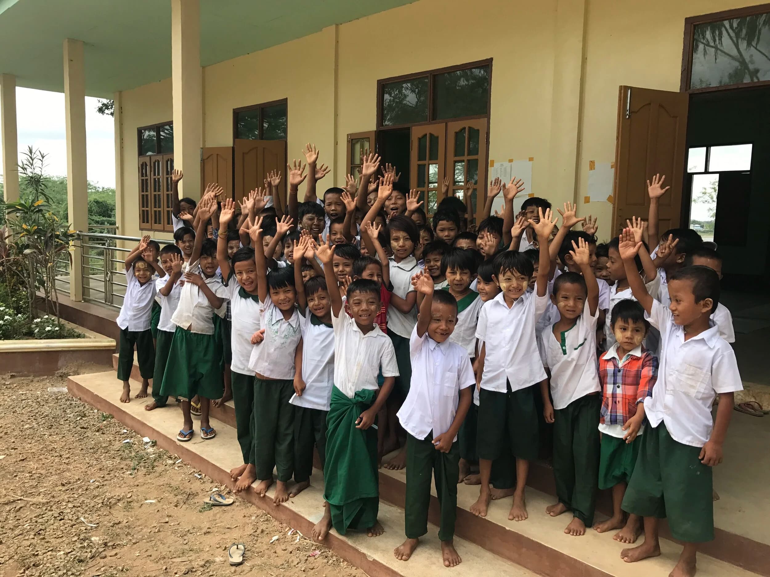 Students in white shirts and green uniforms waving outside their school building