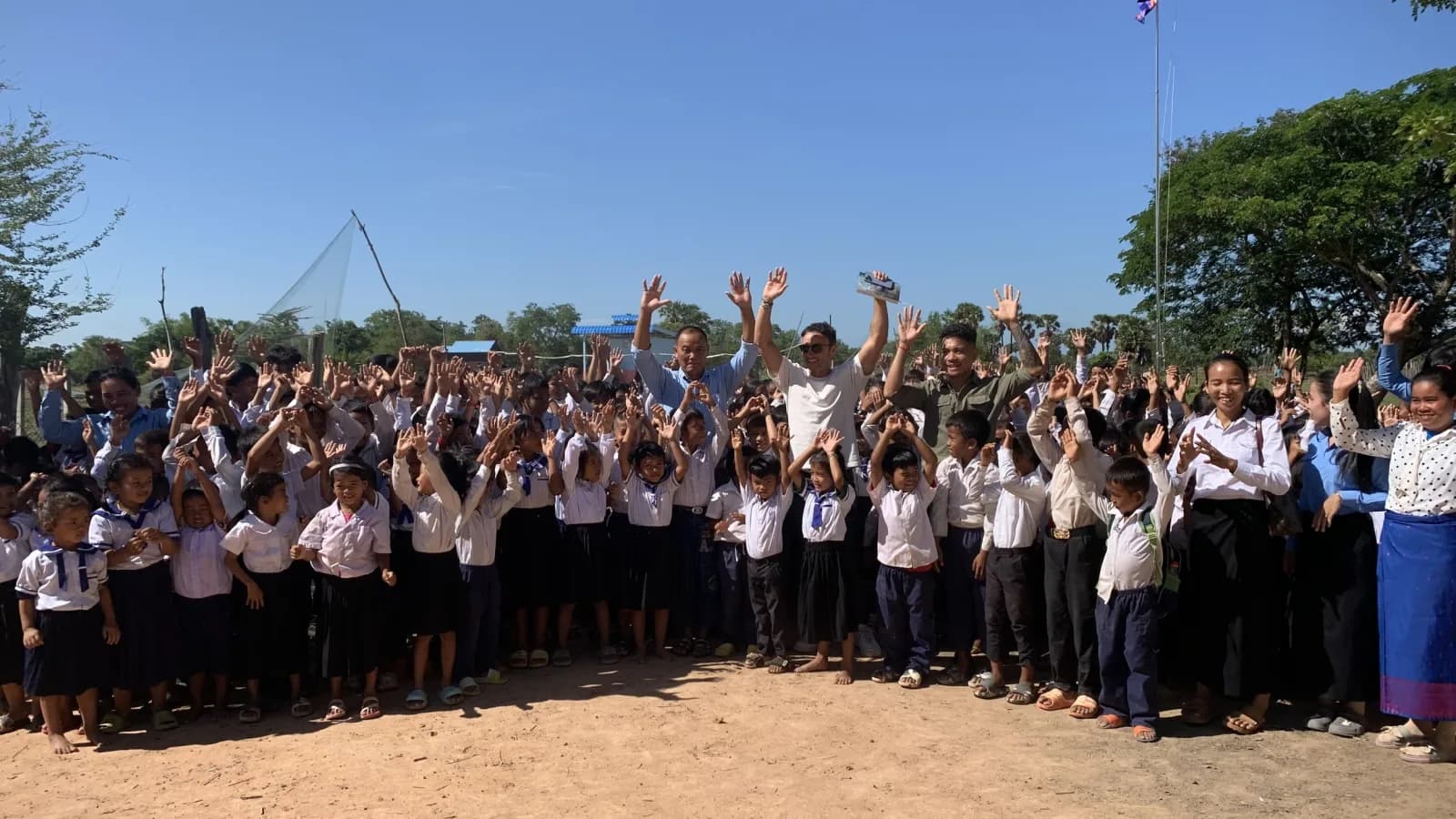 A large group of students and visitors waving their arms in the air outside a school